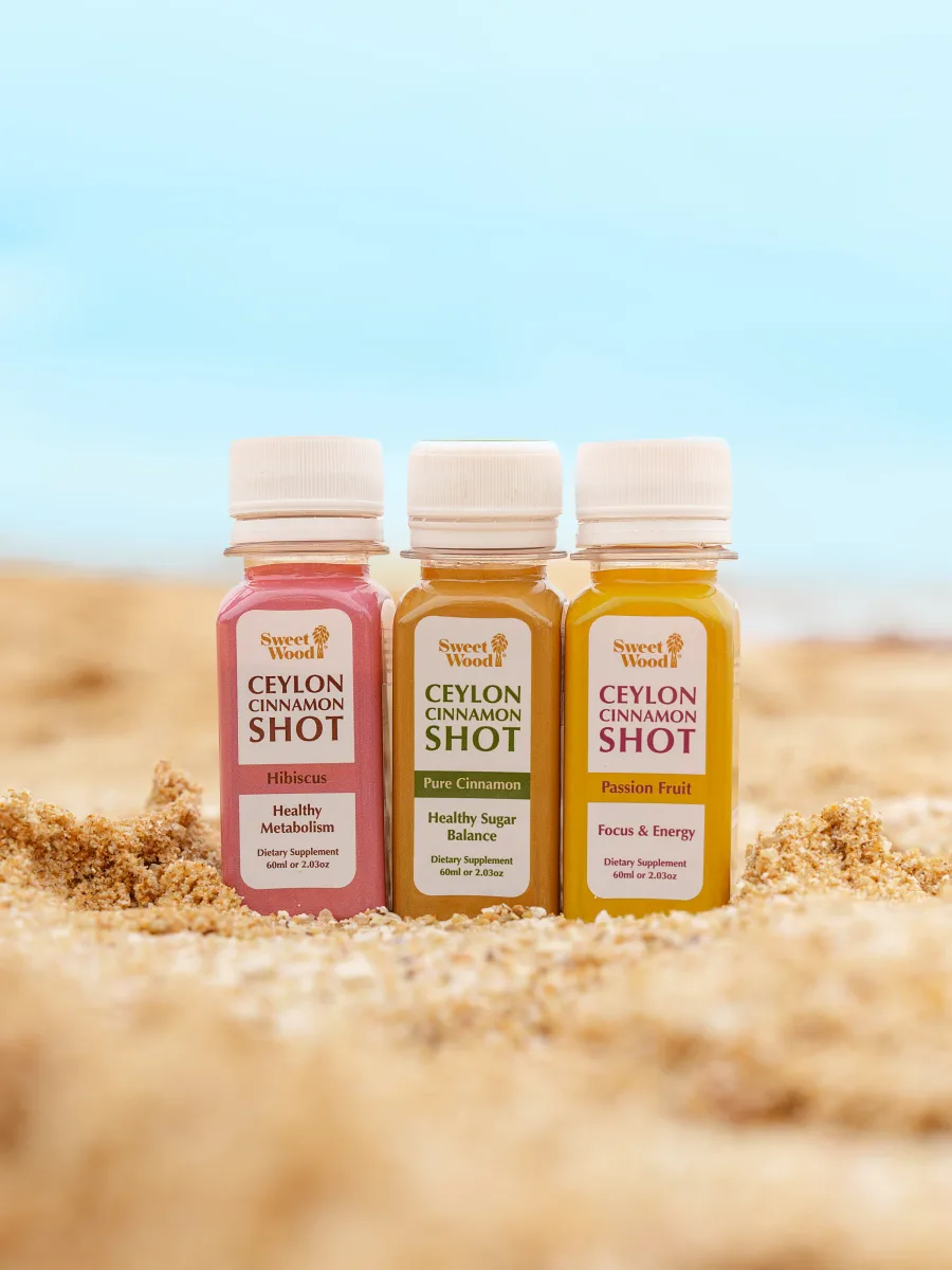 All three Sweet Wood Ceylon Cinnamon Shot flavours with plain white caps standing in beach sand — Hibiscus, Pure Cinnamon and Passion Fruit