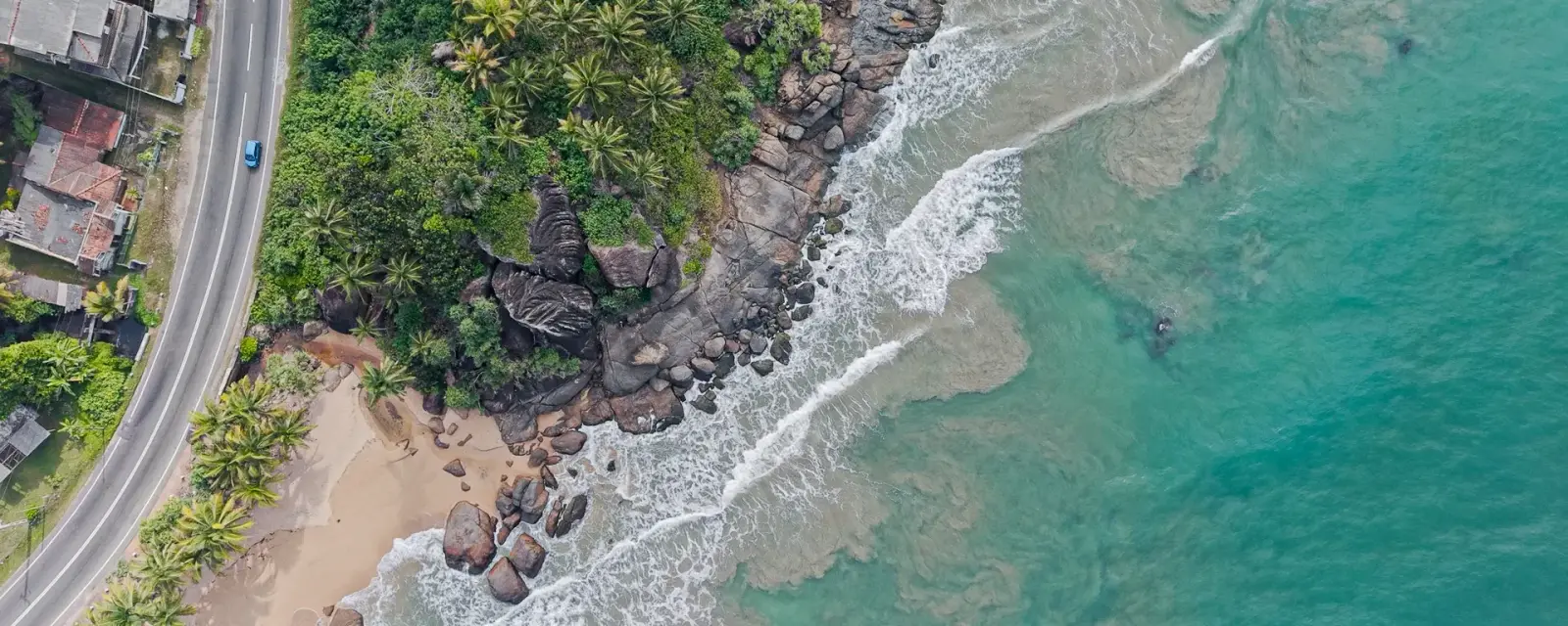 Aerial view of a rocky tropical coastline, with surf breaking against the shore and a coast road running behind the palms